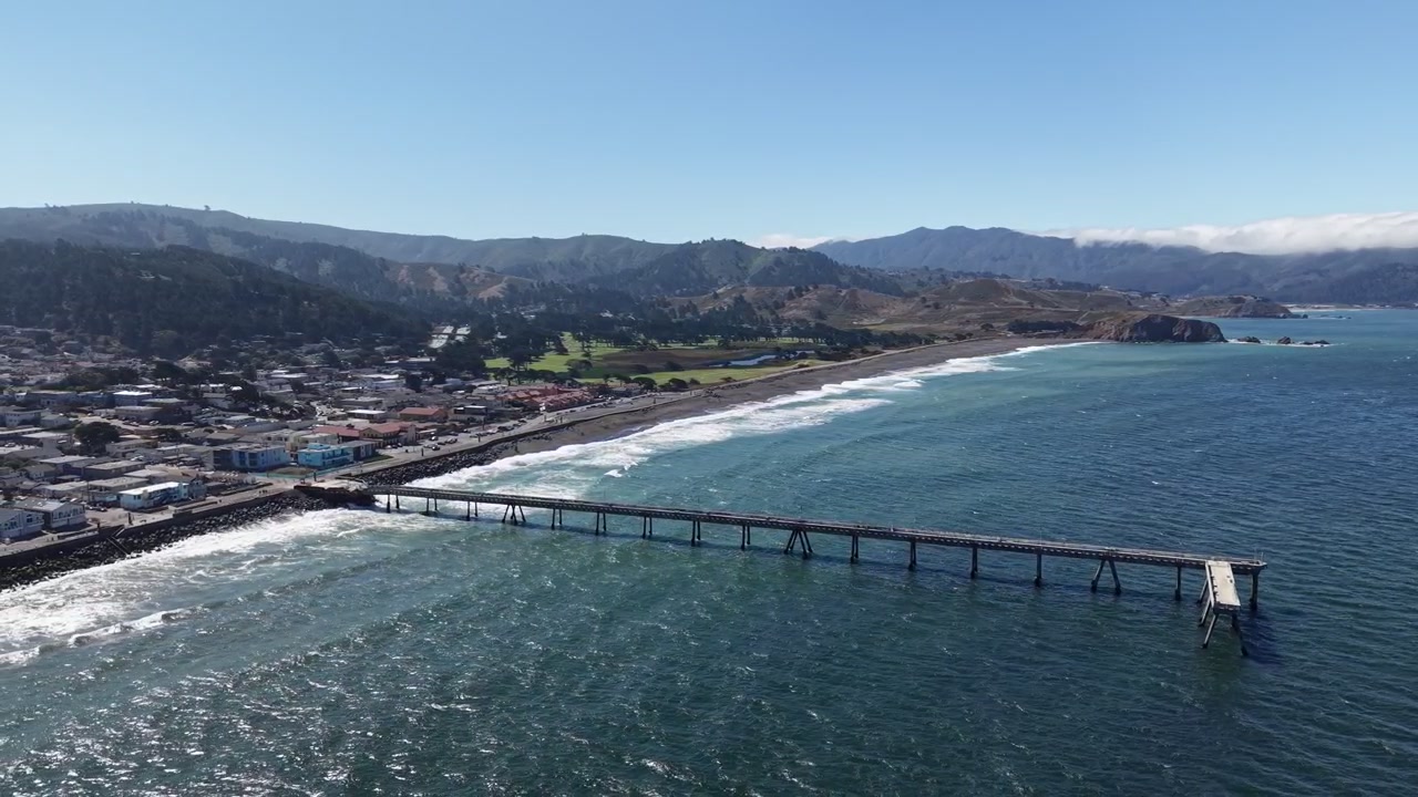 Riprese aeree con drone che sorvolano il Molo Municipale di Pacifica e la scenografica costa della California, mostrando la città, la spiaggia sabbiosa e le dolci colline sotto un cielo azzurro e sereno in una giornata di sole