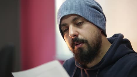 young bearded stylish man with beanie hat reading proposal of his office colleagues, approving new idea by shaking his head suspiciously