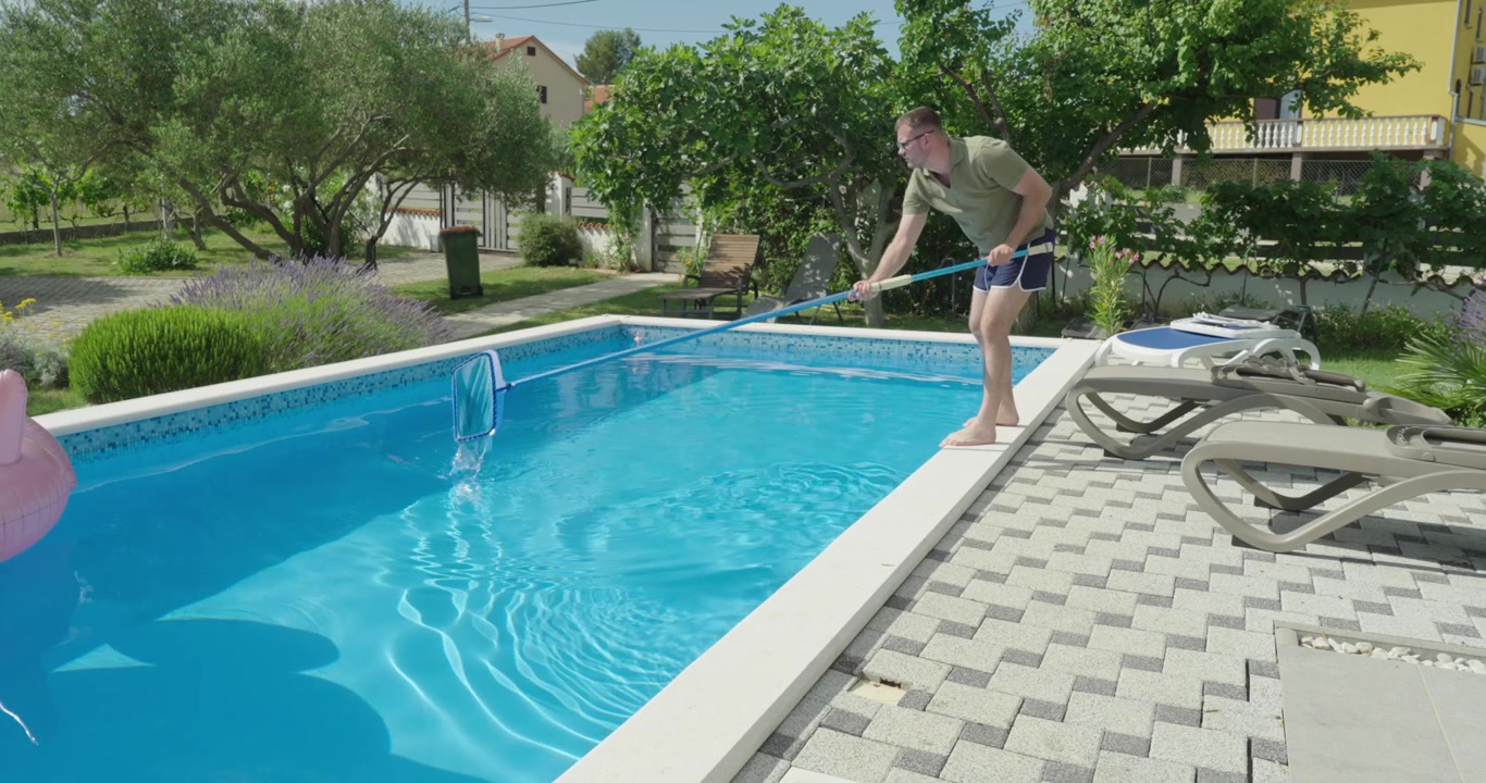 En mand bruger et net til at rengøre en swimmingpool ved at fjerne snavs og uønskede genstande fra vandet. Fokus på poolvedligeholdelse, renlighed og udendørs pleje.