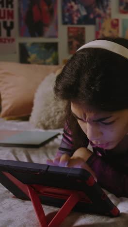 Beautiful-teen-in-headphones-lies-on-bed-in-her-room-and-uses-digital-tablet-computer.-African-American-girl-looks-at-tablet-display-and-makes-funny-faces.-Vertical-shot