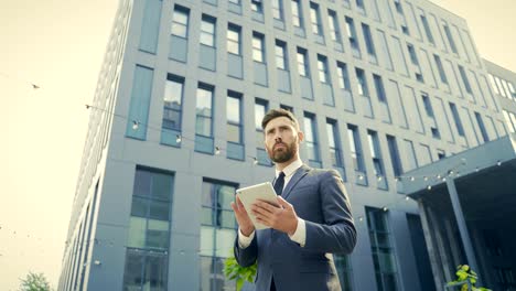 stylish bearded businessman in formal business suit standing working with tablet in hands on background modern office building outside.