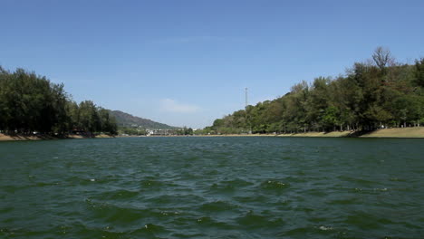 locked low angle shot of panoramic view of nai harn lake on a breezy blue sky summer day, no people, water in foreground and idyllic park and forest in background