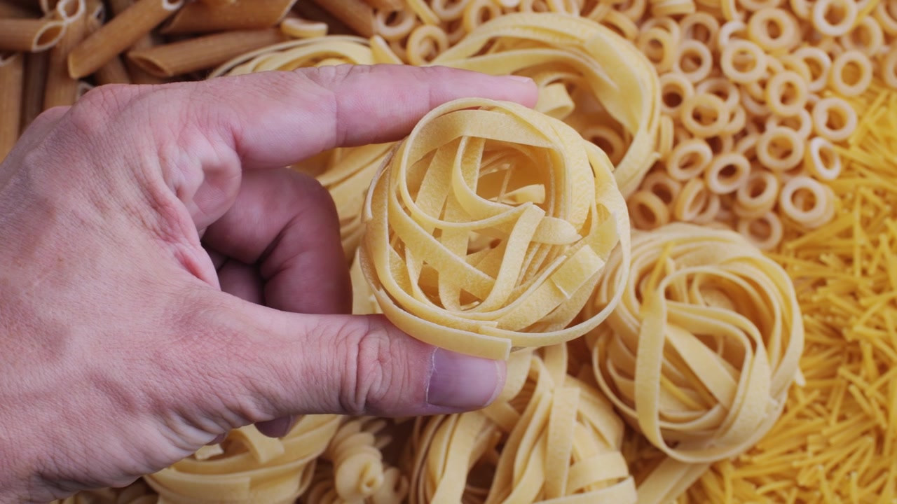 Mano che regge tagliatelle crude con pasta secca assortita sullo sfondo
