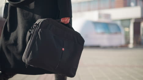 primer plano de una bolsa negra llevada por una mujer con un elegante abrigo negro y botas de cuero caminando con confianza por una calle urbana soleada, con arquitectura moderna borrosa y bicicletas verdes en el fondo