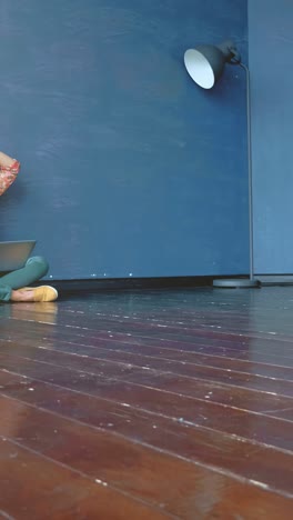 woman with curly red hair is sitting on wooden floor, looking frustrated while working on laptop against a blue wall, conveying a sense of stress and challenge