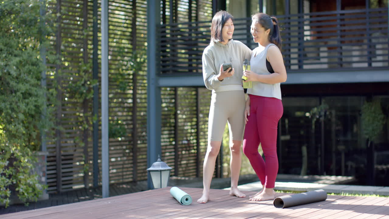 amigas asiáticas felices con botella de agua y teléfono inteligente riendo en la terraza, cámara lenta