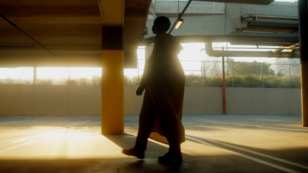 Side profile silhouette of adult in long coat walking across sunlit multi level parking garage, with trailing hem, forward stride, concrete columns, harsh backlight and moody urban atmosphere
