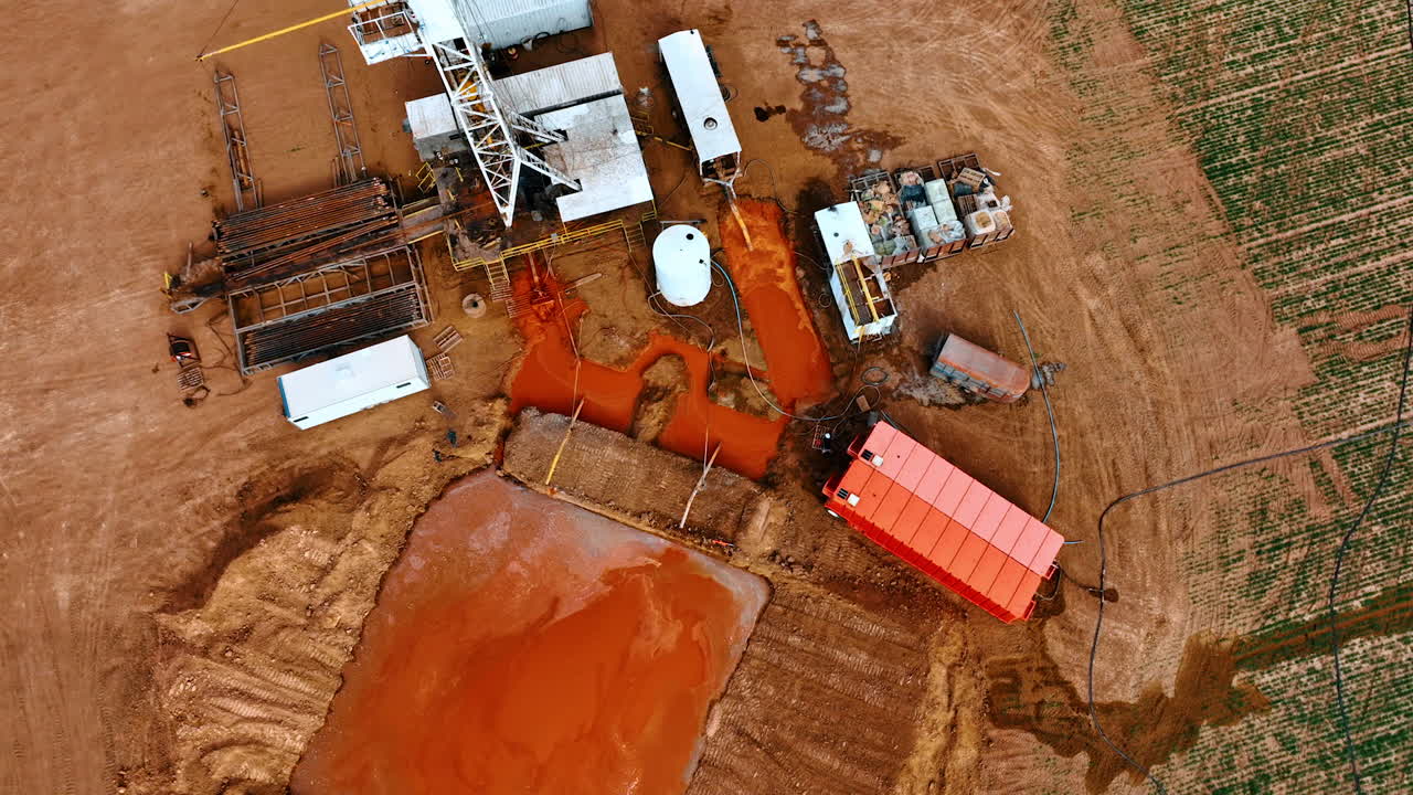 Big square pit filled with orange dirt at the site for drilling oil. Drone footage above the oil rig in the field.