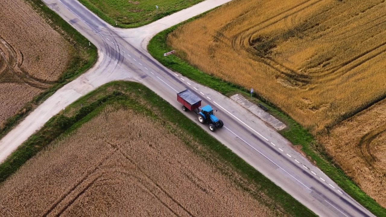 Vista aerea di trattore blu che traina un rimorchio lungo un incrocio tra campi agricoli