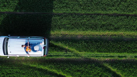 Young man with a guitar on a roof of a car. Hipster guy playing the guitar on the top of a moving car on green field background. Top aerial view.