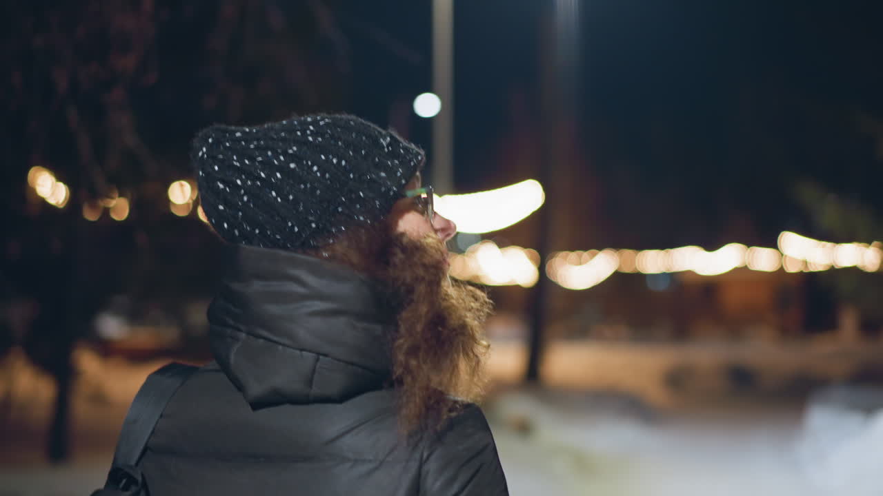 Woman in winter coat and knit hat seen from side walking outdoors at night with snow and glowing festive lights in background, enjoying peaceful cold evening atmosphere illuminated by street lamps