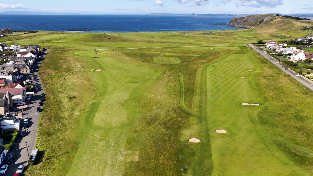 Drone glides above green golf course, seaside village, and coastline under bright daylight