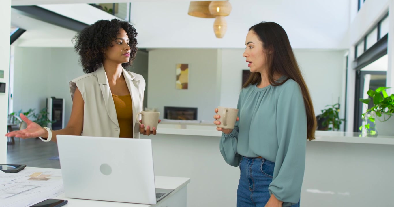 Diverse women in kitchen pointing at plans, leading peer moving white laptop and choosing samples