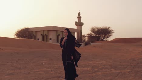 mujer musulmana de pie cerca de la mezquita en el desierto. viento fuerte oriente medio paz sin guerra