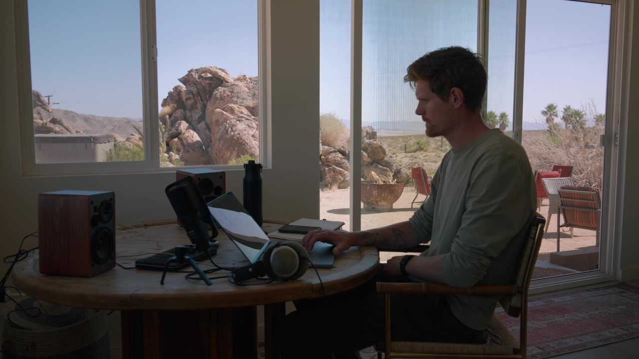 Un uomo alla sua scrivania si toglie le cuffie e sorride, con laptop e altoparlante vicini, e una vista del deserto all'esterno.