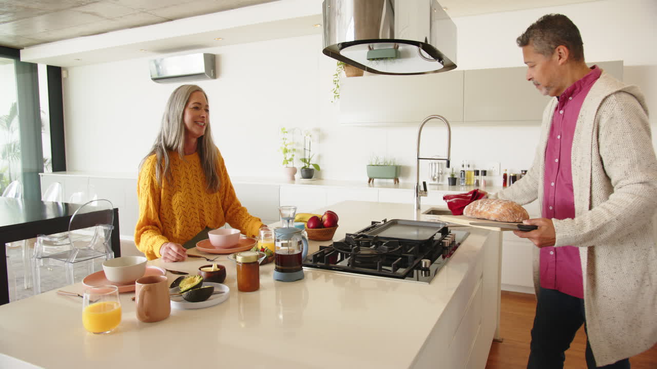 Multiracial senior couple, preparing breakfast, serving bread, smiling in modern kitchen, at home