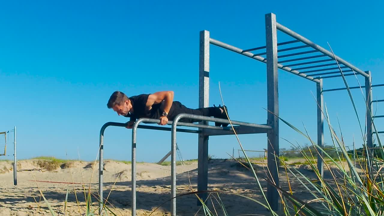 Beautiful static footage of a man with tattoos working out and doing pushup exercise in an outdoor metal gym at a beach during sunny warm and hot day. Man is fit and muscles are streched during pushes
