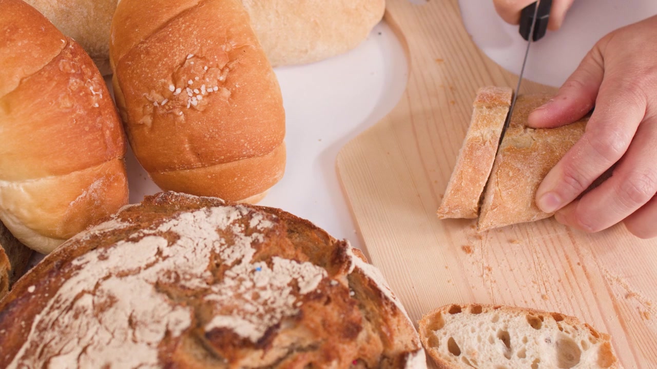 Primo piano di mani che affettano pane croccante su un tagliere di legno tra assortimenti di prodotti da forno.