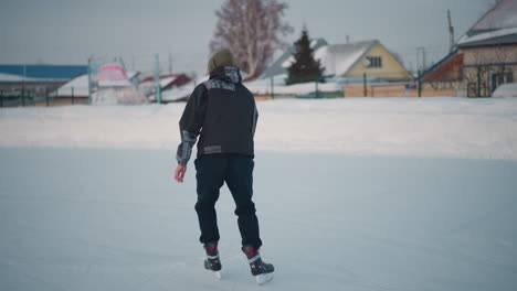 back view of boy gliding smoothly on icy suburban rink under pale sky, patterned jacket and green hoodie visible, distant blur of solitary figure near snowy banks