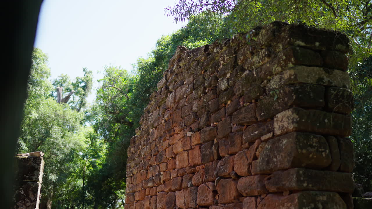 Sunlight illuminating a weathered red sandstone wall of the Jesuit reduction San Ignacio Miní, a UNESCO World Heritage site, surrounded by lush vegetation in Misiones, Argentina, slow motion reveal