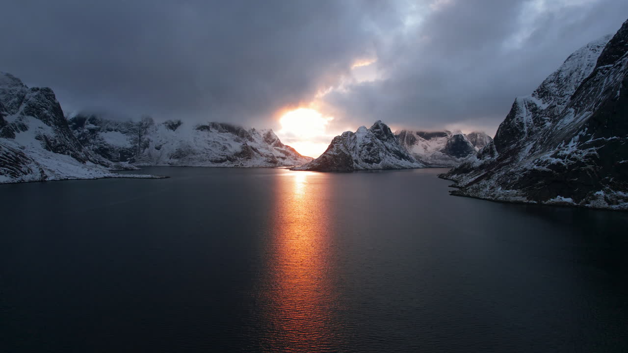 Stunning drone flight over Reine, Lofoten Islands, Norway, with the sun peeking through clouds over the water and snowy mountains