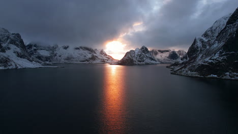 Stunning-drone-flight-over-Reine,-Lofoten-Islands,-Norway,-with-the-sun-peeking-through-clouds-over-the-water-and-snowy-mountains