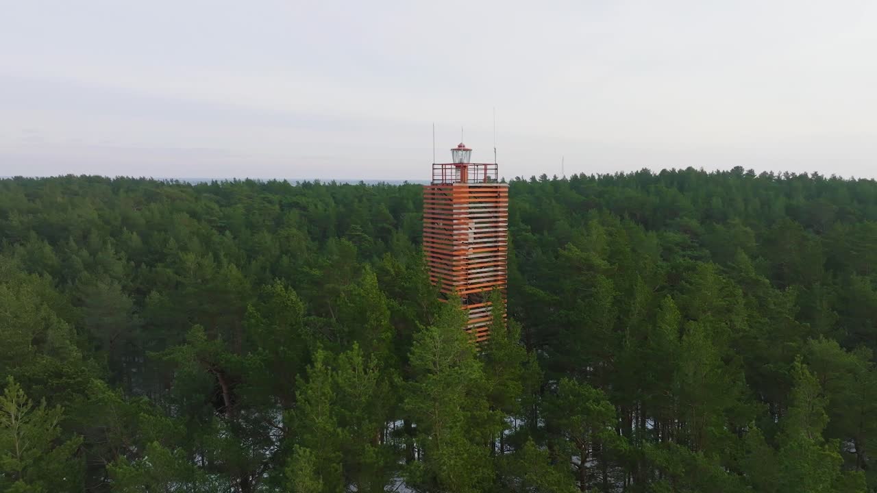 Aerial view of Bernati lighthouse surrounded by lush green pine tree forest with light snow, Nordic woodland, Baltic sea coast, sunny winter day, Latvia, wide ascending drone shot moving forward