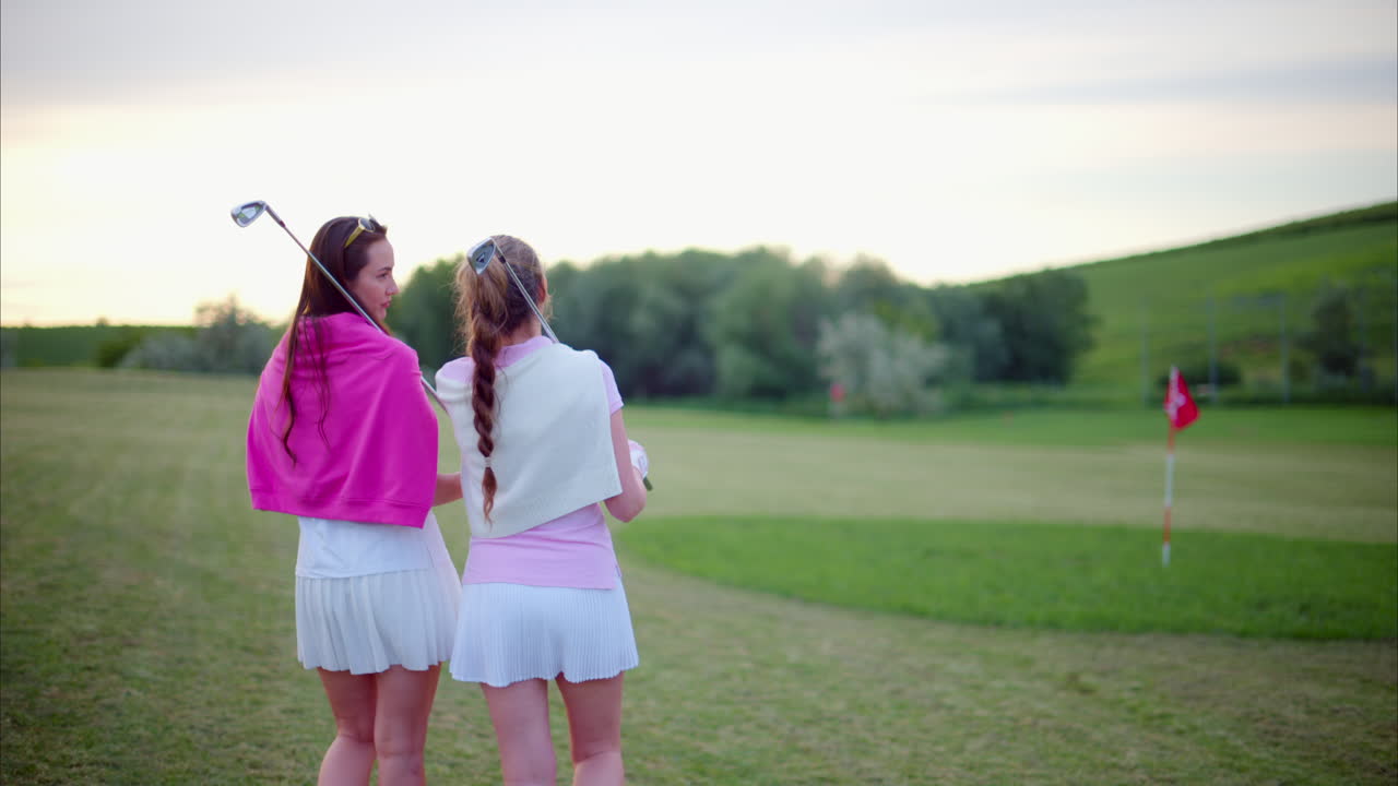 Two women dressed in white and pink clothes, with golf clubs on their shoulders, walking and talking on the golf course