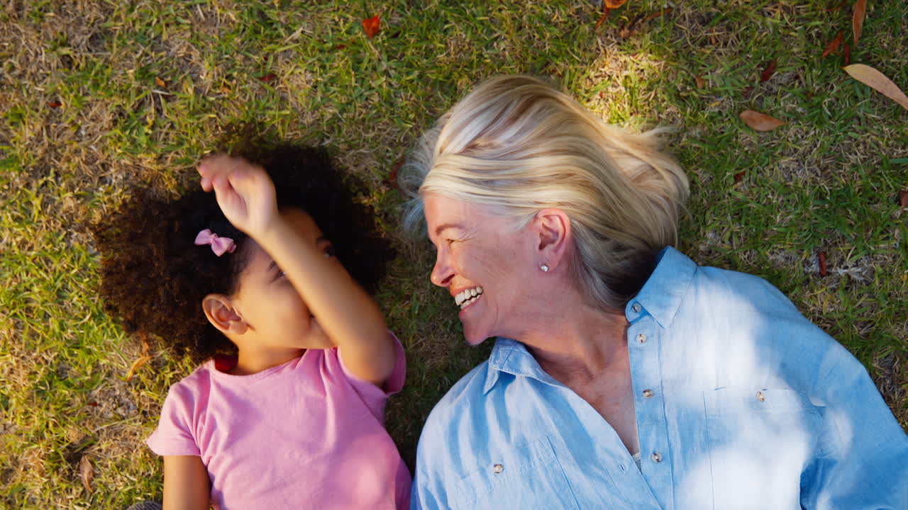 Overhead Shot Of Grandmother And Granddaughter Lying On Grass Talking Together