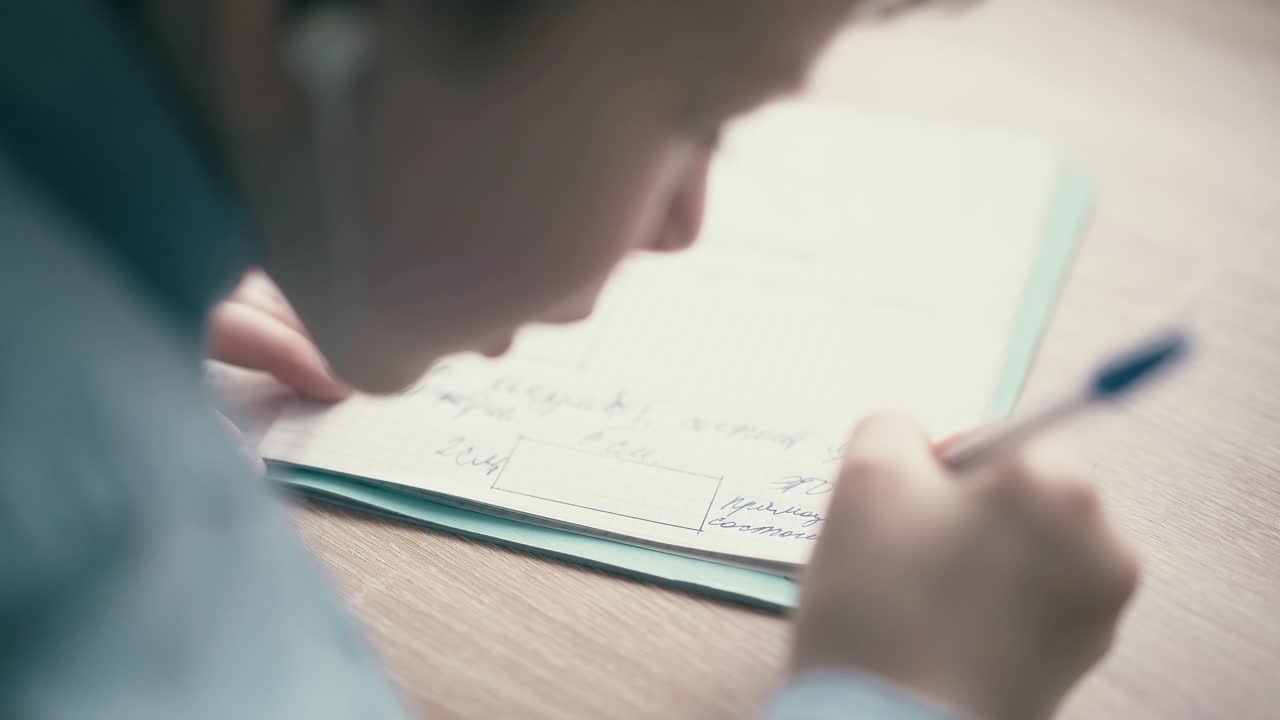 cu 소년은 헤드폰에 앉아서 학교 숙제를하고 노트북에 볼펜을 쓰고 있습니다.