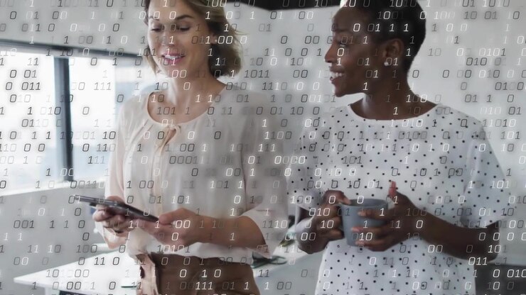 Discussing two coworkers in cream blouse and polka-dot blouse using tablet, holding mug in office