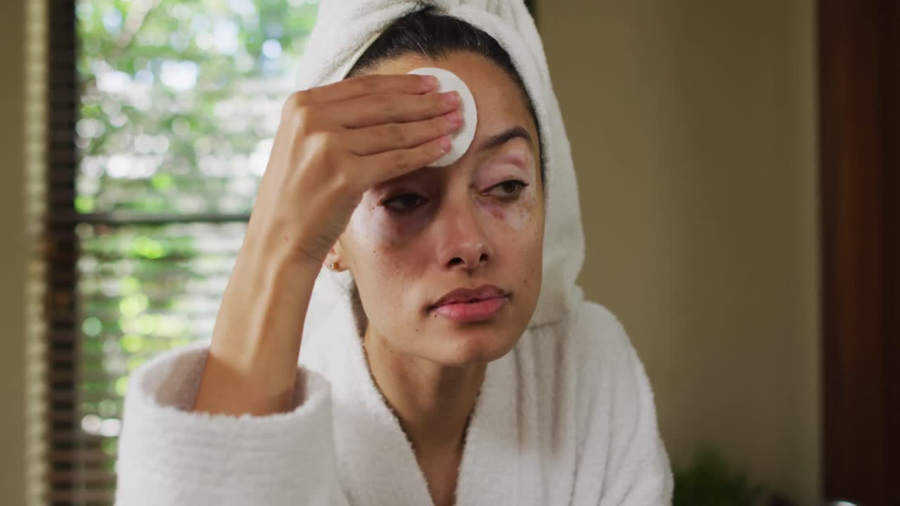 Portrait of biracial woman in robe looking into mirror and cleansing face