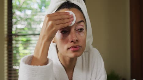 portrait of biracial woman in robe looking into mirror and cleansing face