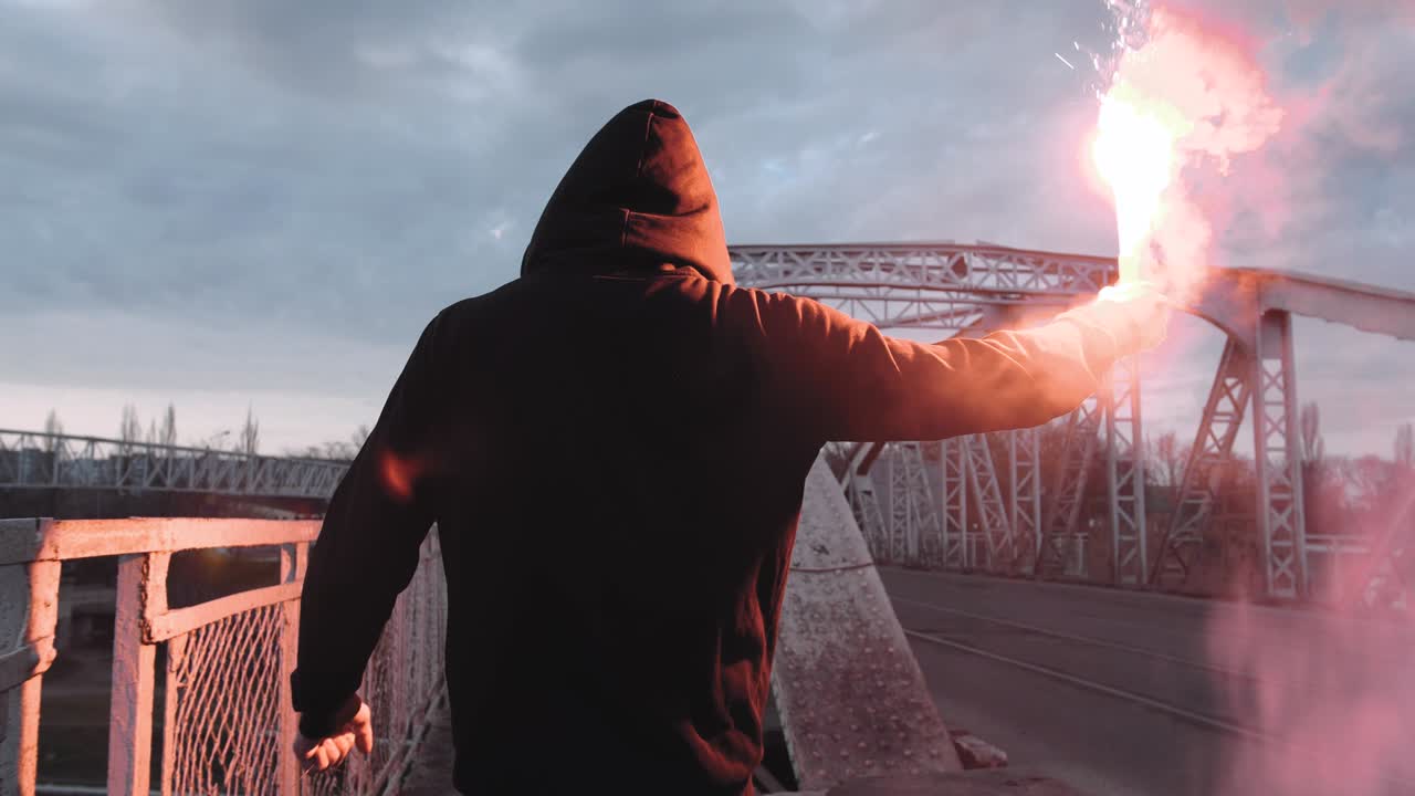 задний вид молодого человека в капюшоне и балаклаве с красной горящей сигнальной факелой на дороге под старым стальным мостом, замедленное движение