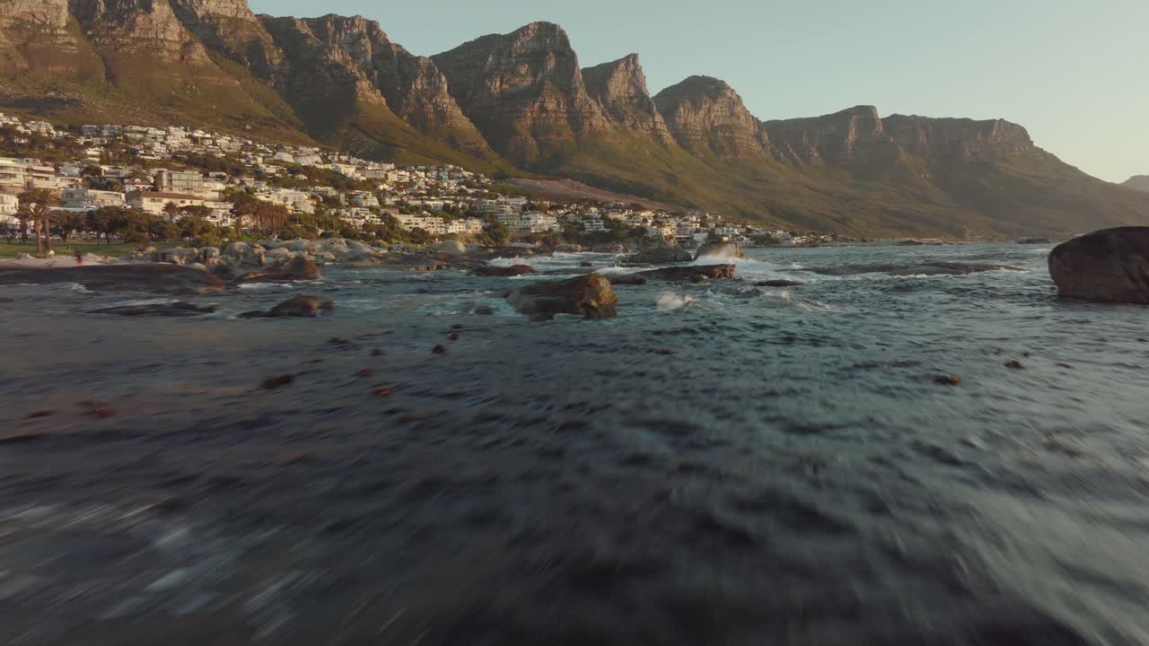 Drone flies just over the sea at camps bay in cape town south africa - wavy sea, many houses on a hilltop - drone flies with view to table mountain rocks in the sea