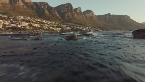 drone flies just over the sea at camps bay in cape town south africa - wavy sea, many houses on a hilltop - drone flies with view to table mountain rocks in the sea