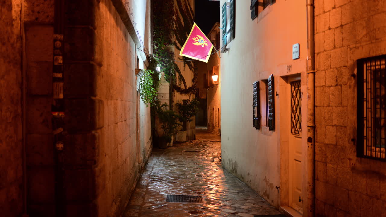Bandiera del Montenegro appesa in un tradizionale vicolo stretto in pietra con strada acciottolata e calda illuminazione notturna. Città Vecchia di Cattaro
