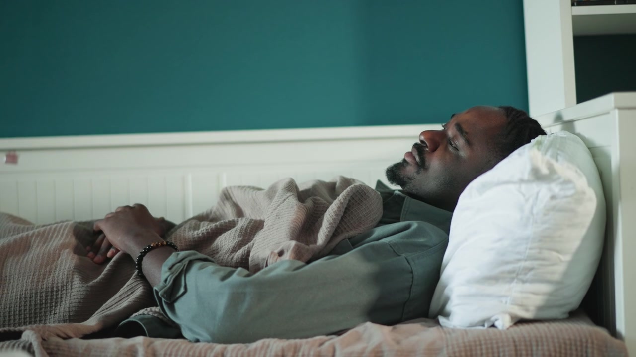 Black man lying in bed, teal bedroom, wrapped in knit blanket, pillow supporting head, eyes closed in quiet recovery after emergency, calm expression, subtle sunlight, intimate closeup, home health