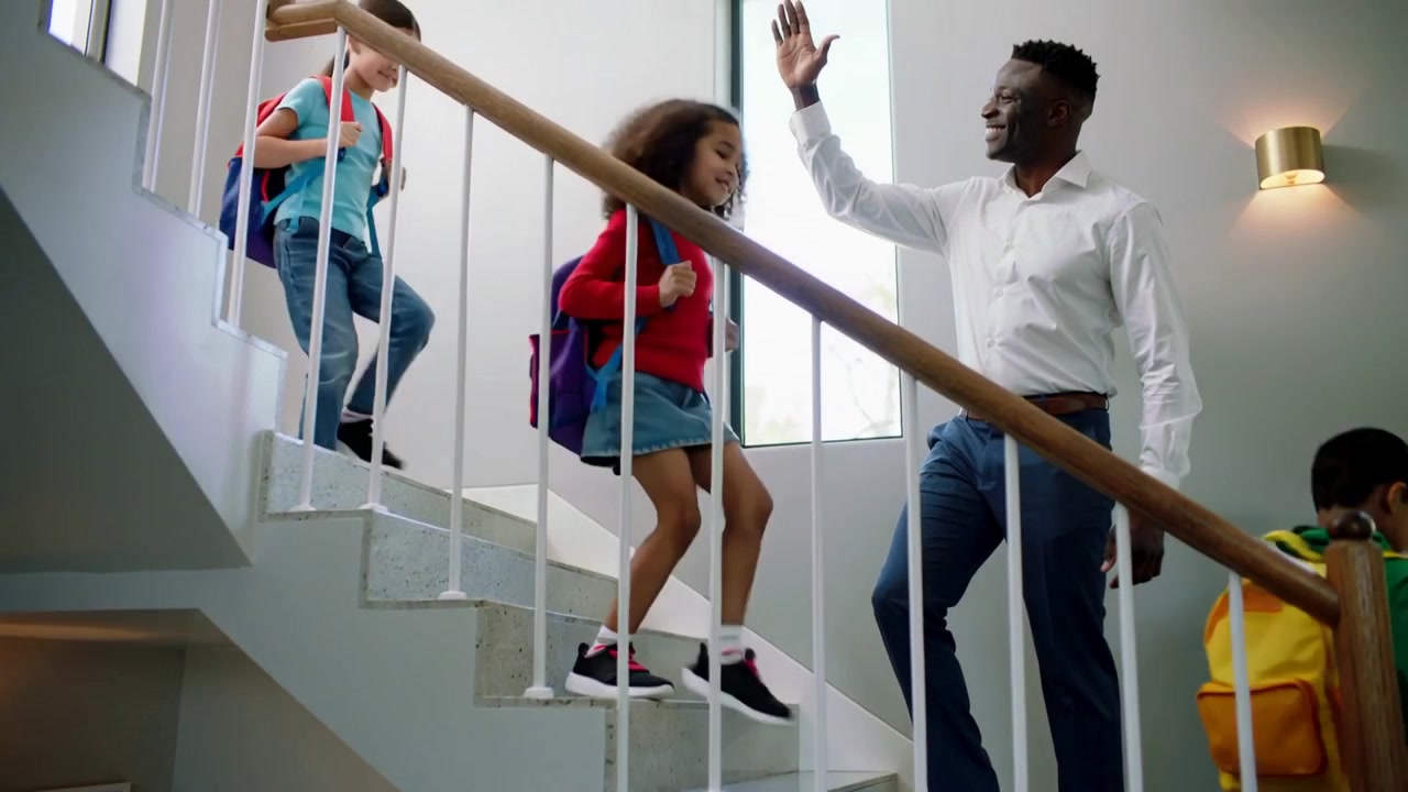 En smilende mand i hvid skjorte løfter hånden og hilser på børn, der forlader trapperne til skolen med rygsække.