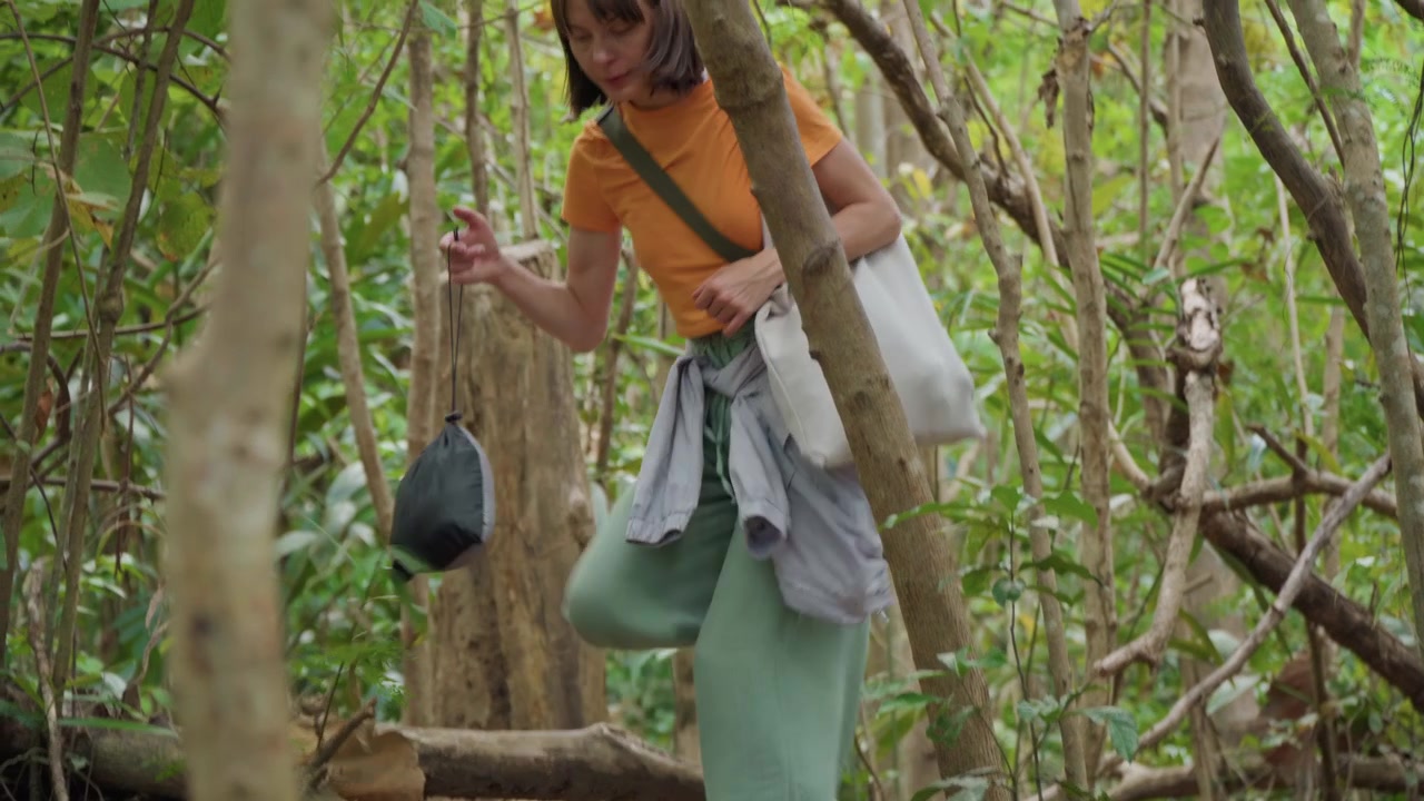 Close up of woman walking through dense forest with green foliage and tree trunks carrying bags and wearing casual outdoor clothing. Woman navigates narrow paths between plants while holding