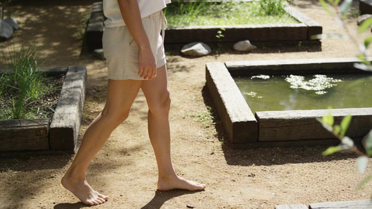 裸足の彼女が、明るいショートパンツとトップス姿で、池と植木鉢のそばを通りながら庭の小道を穏やかに進む。