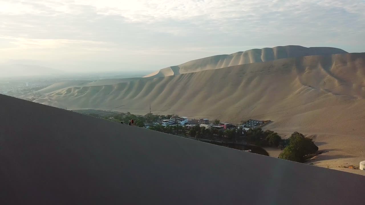 aéreo, seguimiento, disparo de drones, de un hombre y una mujer sentados en una duna, mirando la ciudad del oasis de huacachina, al atardecer, en una tarde soleada, en el suroeste de perú