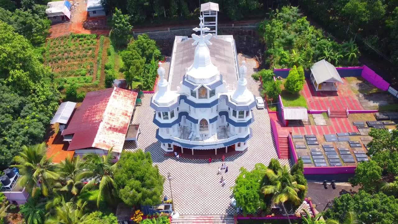 High-angle drone footage descending over a beautiful Syro-Malabar church and its cemetery, nestled amongst lush tropical foliage and farmland in the Keralan countryside, India, on a bright sunny day