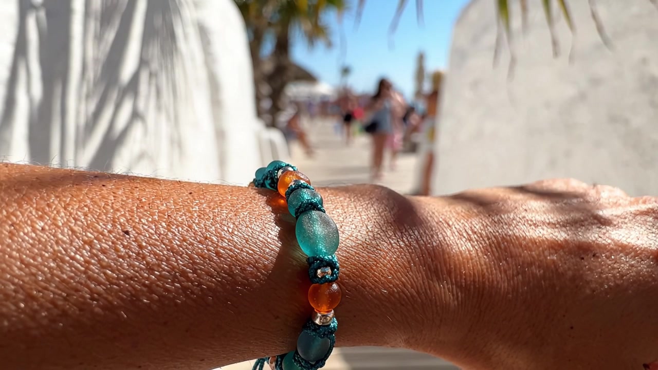 Un bellissimo primo piano di un braccialetto di perline colorato su un braccio baciato dal sole, sullo sfondo di una vivace scena estiva in spiaggia a mezzogiorno.
