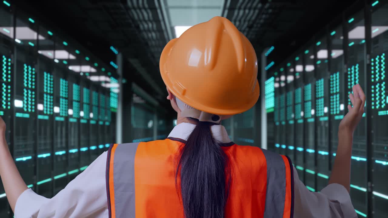 Primo piano da dietro di un'ingegnera con casco di sicurezza che allarga le braccia mentre è in piedi nella sala server di un data center