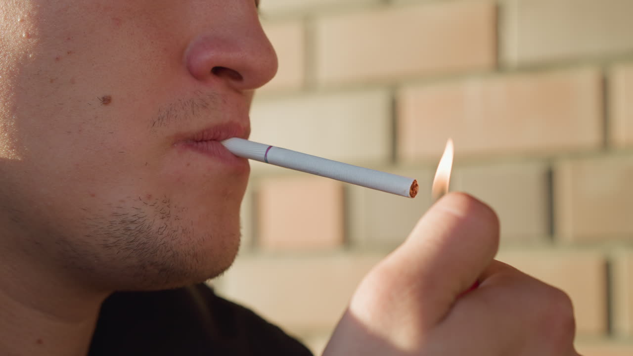 close up of man placing cigarette between lips while holding lighter near cigarette tip, preparing to ignite cigarette against soft daylight and blurred brick wall background