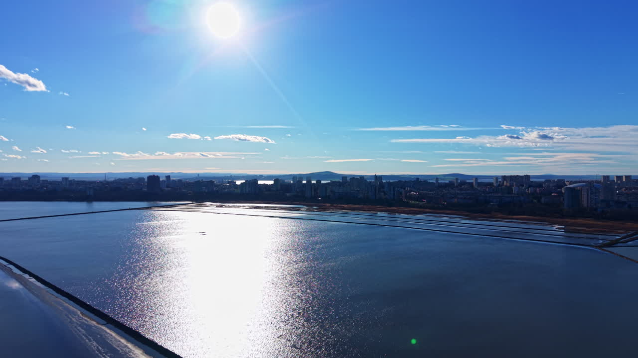 日中の澄み切った青空の下、明るい太陽が街の近くの広大な水面を照らし、遠くには海岸線がはっきりと見える。
