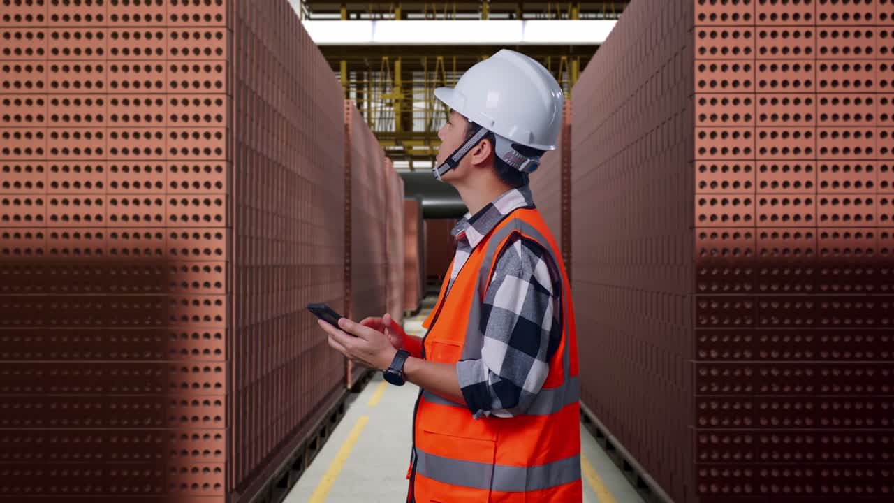 Side View Of Asian Male Engineer With Safety Helmet Using Smartphone And Looking Around While Standing With Red Brick Packed in Stacks Are Stored