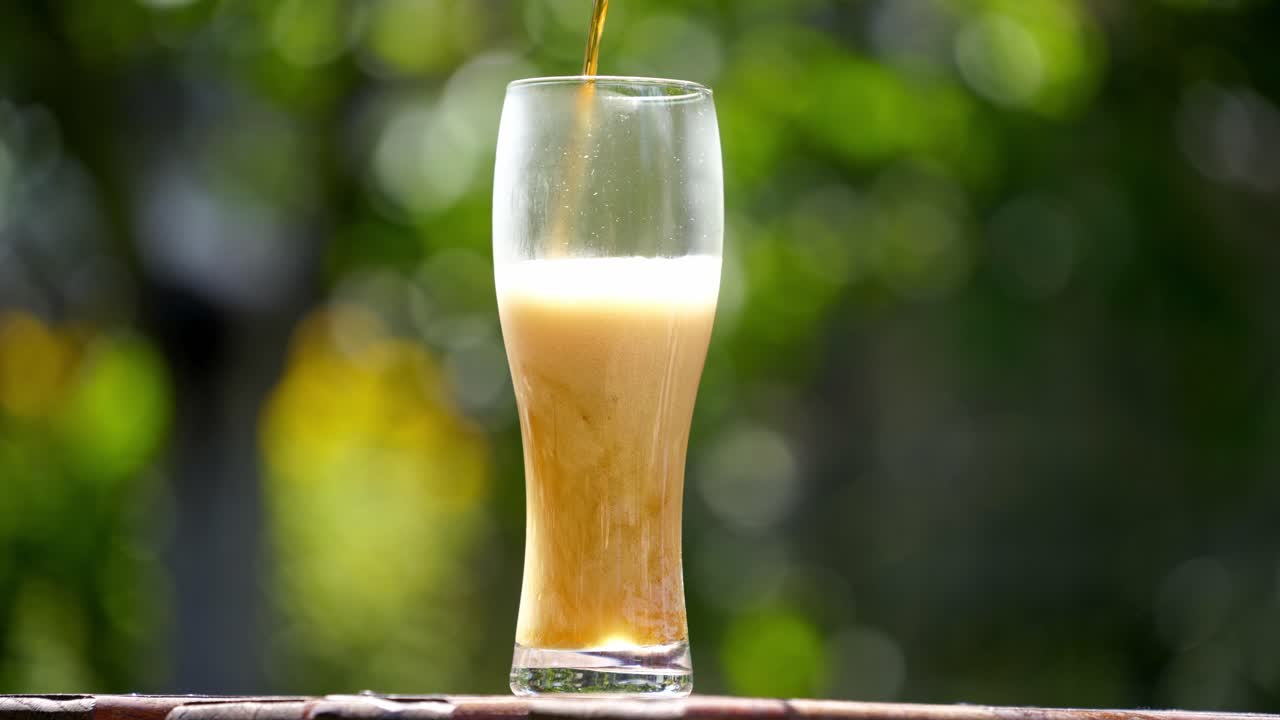 Pouring process of light beer into tall glass. Beer pours into a glass in summer garden making lots of bubbles and white foam. Cold alcohol refreshment on nature background.