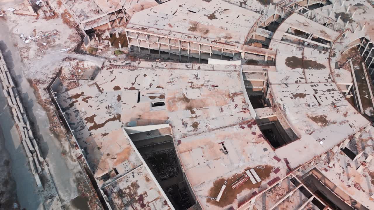Oblique overhead drone of a semi-circular abandoned construction site with exposed beams, cracked slabs, stains and debris—gritty demolition survey urbex visual no people